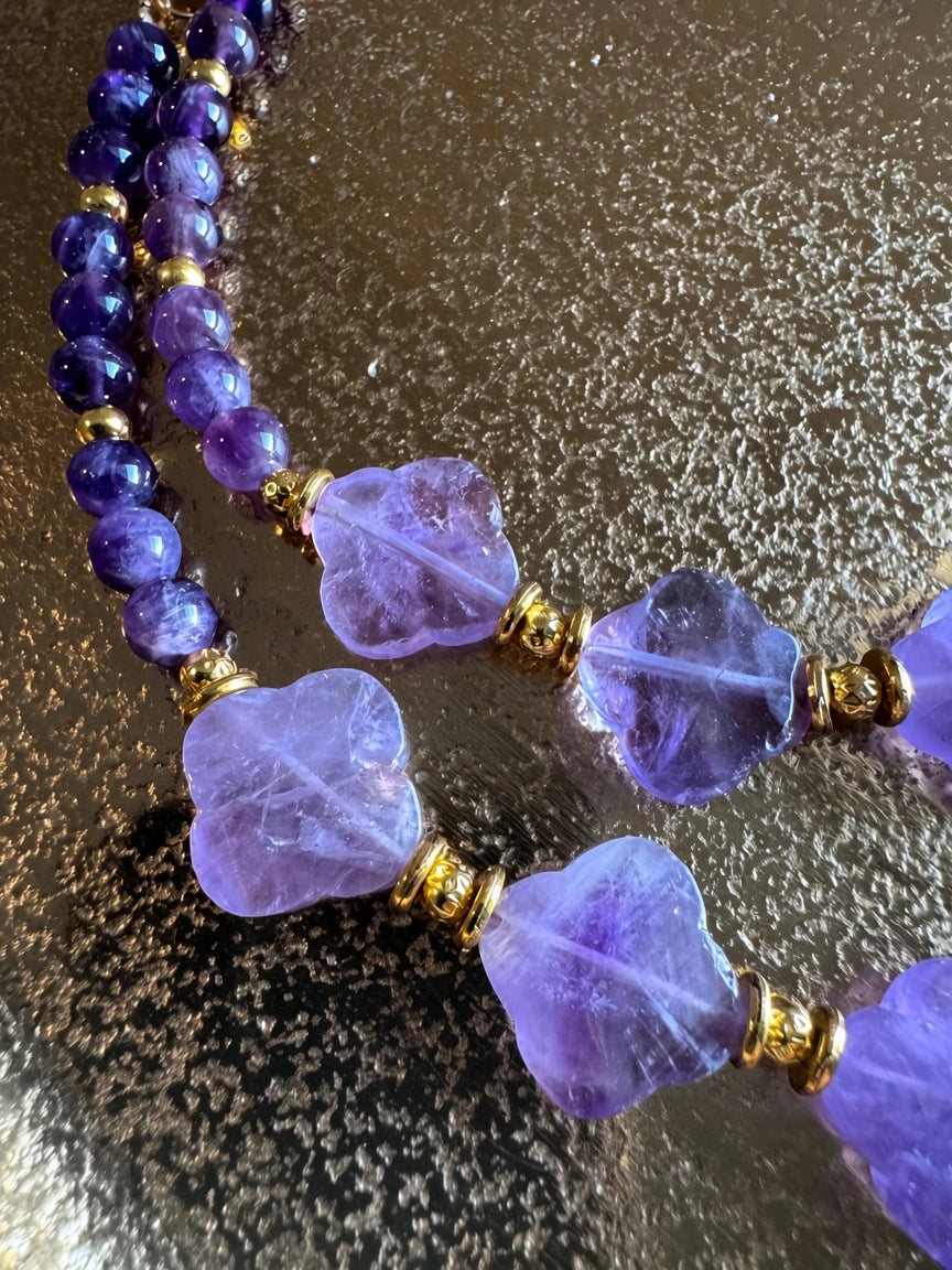 Hue La La Gems amethyst beaded necklace with purple clover-shaped stones and gold-tone spacers on a metallic surface.