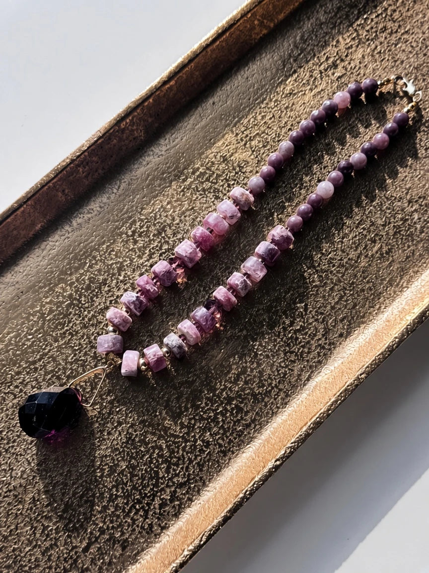 Hue La La Gems beaded statement necklace in plum and mauve with a faceted dark purple crystal pendant on a gold tray.