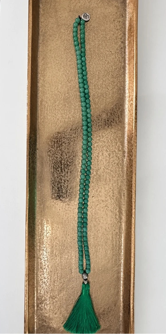 Hue La La Gems green aventurine 108-bead mala necklace with a silver Om charm and deep green silk tassel on a gold tray